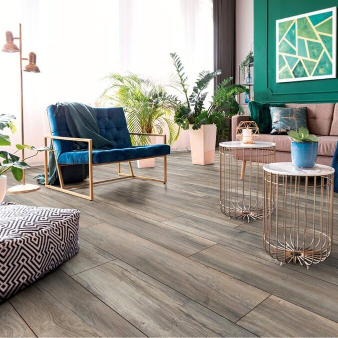 Raumfoto eines Laminats in grauer Holzoptik in einem modernen Wohnzimmer in den Farben Hellbraun, Rosé und Grün.  Im Hintergrund sind Tische und ein Stuhl zu erkennen.