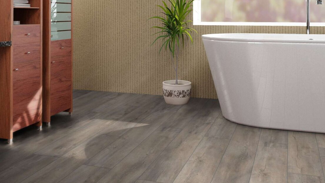 Laminat in grauer Holzoptik in einem Badezimmer. Im Hintergrund sind dunkelbraune Schränke sowie eine moderne weiße Keramikbadewanne.