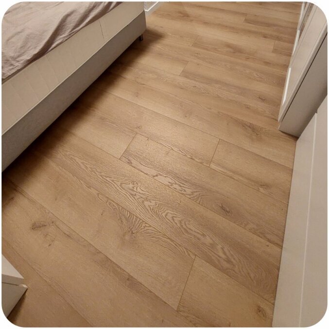 Nahaufnahme eines Klick-Laminatbodens in einem Schlafzimmer. Das Laminat hat eine natürliche hellbraune Farbe und gut sichtbare Maserungen sowie Fugen, was den Eindruck von echtem Holzboden erweckt. 