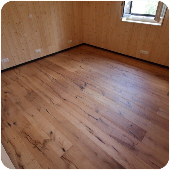 Raumbild eines Parkettbodens. Der Parkettboden hat eine dunkelbraune Farbe und viele Furchen und Risse auf der Oberfläche und damit einen rustikalen Charakter. Die Wände des Raums sind mit etwas hellerem Holz verkleidet und es sind Steckdosen sowie ein Fenster sichtbar. 