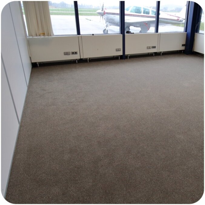 Raumbild eines grauen Teppichs in einem Raum mit vielen hohen Fenstern. Durch die Fenster ist eine Landebahn sowie ein Flugzeug erkennbar. Bei dem Teppichboden handelt es sich nicht um Rollenware sondern um einzelne Teppichfliesen, die miteinander verbunden worden sind und dadurch nicht unterscheidbar von regulärer Rollenware sind. 