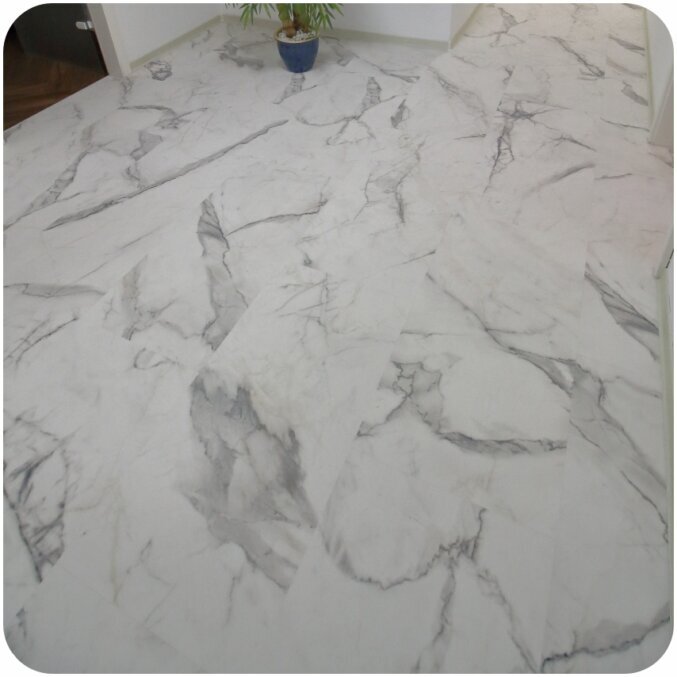 Nahaufnahme eines Flurs bei dem Vinylboden in Fliesenoptik verlegt wurde. Das Vinyl hat die Optik von weißen Fliesen mit grauen Marmorelementen. Die Sockelleisten sind ebenfalls in Weiß gehalten.