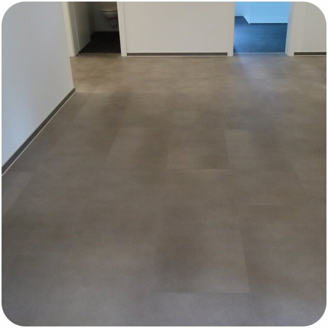 Raumbild eines breiten Flurs. Erkennbar ist ein Vinylboden in schlichter und dunkelgrauer Fliesenoptik. Der Boden erinnert an Steinfliesen und hat so ein dezentes Muster, dass er fast Einfarbig wirkt. Es sind außerdem Sockelleisten zu erkennen, die einen etwas dunkleren Grauton haben und so einen Kontrast zum Boden und zur weißen Wand bieten.