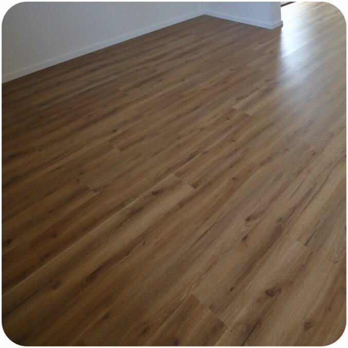 Aufnahme eines Raums in dem ein Vinylboden liegt. Die Optik des Bodens ist in einem dunkelbraun gehalten und hat leiche Furchen und Risse. Dadurch gibt der Boden eine nur leicht rustikale Ausstrahlung ab. Die Sockelleisten sind passend zu den Wänden in der Farbe Weiß.